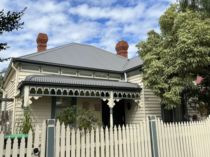 Close-up image of a white house in Woodland Grey Brunswick, featuring a gray Colorbond roof with a chimney, a bullnose veranda, a white fence, and surrounding trees.